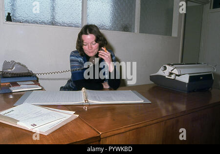 1970s, historical, a female office worker, perhaps an accounts clerk, sitting at her desk looking at numbers in a clip file speaking on the telephone, England, UK. Stock Photo