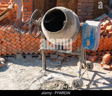 close up of concrete mixer at dirty construction site near packs pallets of red bricks. summer noon, outside shot Stock Photo