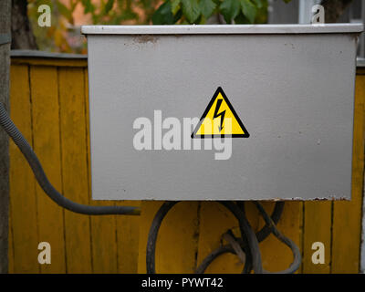 close up of closed gray electrical junction box with caution warning danger sign thunder in yellow triangle. wires sticking out of the box. outdoor sh Stock Photo