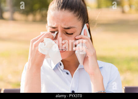 Sad young woman cries while talking on the mobile phone in the park on bench Stock Photo