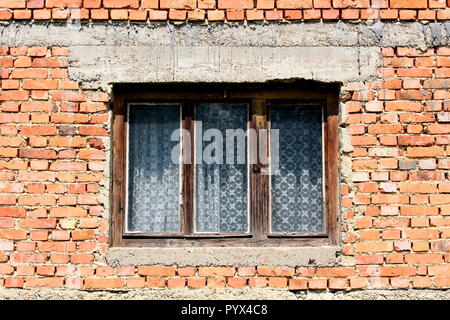 Old dilapidated house window with white curtain and cracked faded wooden frame on red brick wall on warm sunny day Stock Photo