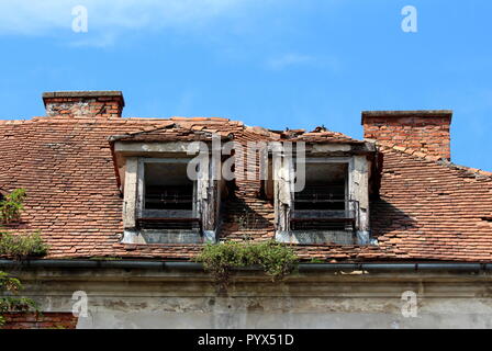 Two destroyed roof windows with rusted metal flower holders on abandoned old building with broken and missing roof tiles surrounded with rusted gutter Stock Photo