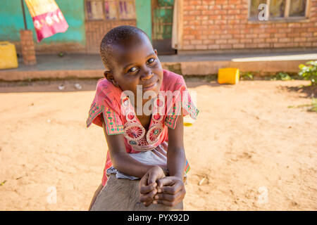 A beautiful young Ugandan black girl leaning on a wooden bench. She has a serene yet happy look. She is dressed a pink ornamented T-shirt. Stock Photo