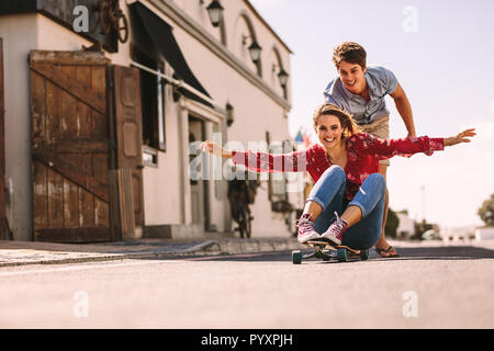 Smiling woman sitting on a skateboard sliding down an empty street. Man helping his girlfriend ride on a skateboard pushing her from behind. Stock Photo