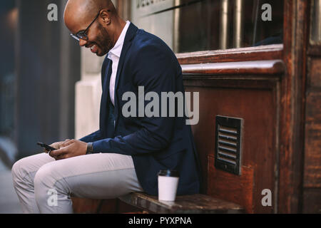 Smiling young man sitting on wooden bench outdoors and reading text message in his mobile phone. businessman using smart phone sitting outside. Stock Photo