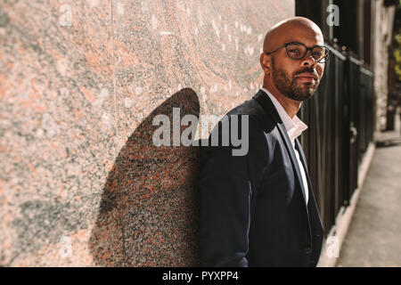 Portrait of handsome young man wearing suit leaning to a wall outdoors and looking at camera. African businessman with beard wearing eyeglasses relaxi Stock Photo