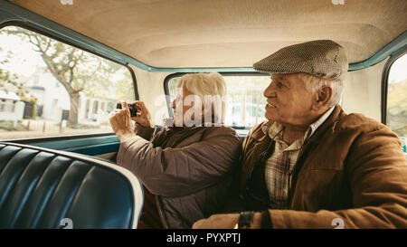 Senior man sitting in backseat of car with wife taking pictures with digital camera. Old couple taking photographs on road trip. Stock Photo