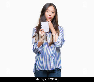 Young asian woman holding notebook over isolated background serious face thinking about question, very confused idea Stock Photo