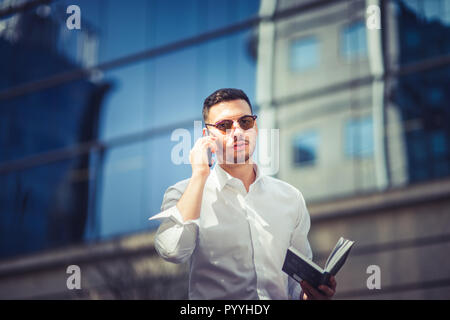 Urban businessman is making a phone call while walking near buildings with glasses, with sunglasses on and notebook in his hands Stock Photo