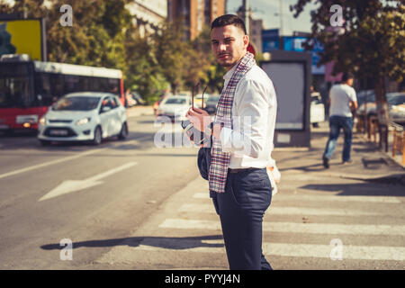 A modern businessman is looking curious in the city while crossing the pedestrian with coffee and mobile in his hands Stock Photo