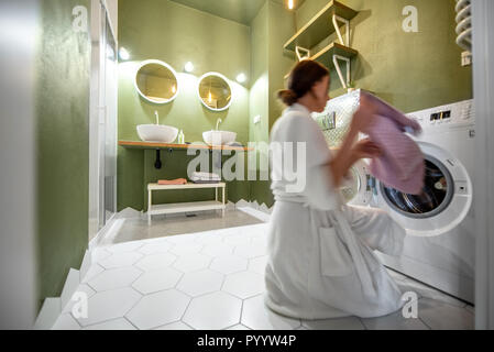 Woman in bathrobe taking off clothes from the washing machine in the bathroom. Woman is motion blurred Stock Photo