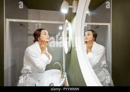 Woman in bathrobe making facial massage looking into the round mirror in the bathroom Stock Photo