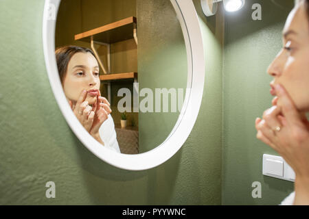 Woman in bathrobe making facial massage looking into the round mirror in the bathroom Stock Photo