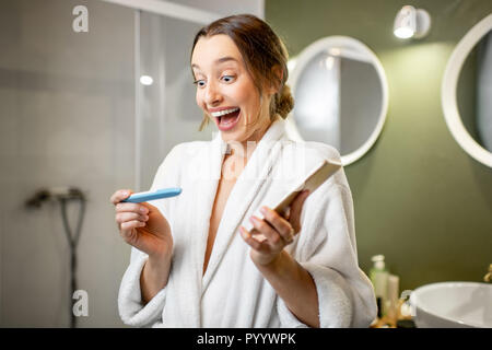 Happy woman in bathrobe excited with a pregnancy test result in the bathroom Stock Photo