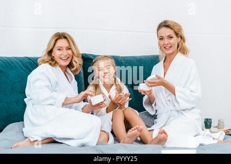laughing three generations of women in bathrobes sitting on bed at home and applying moisturizing cream Stock Photo