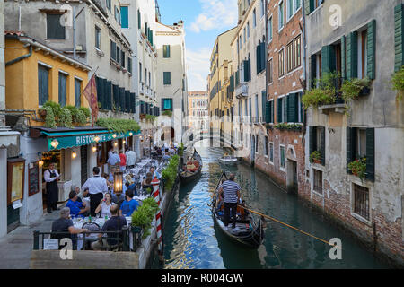 VENICE, ITALY - AUGUST 13, 2017: Restaurant with sidewalk tables with people at dinner and gondola passing in the canal Stock Photo