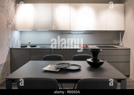 Compact modern grey and white fitted kitchen with illuminated cabinets and a dining table set with ceramics Stock Photo