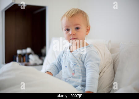 Little toddler boy in pajama with running nose and fever, lying in bed, mom taking care of him at home