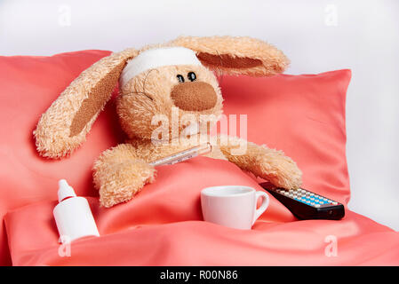 A toy unhealthy rabbit with a thermometer and a TV remote control is lying in bed. Stock Photo