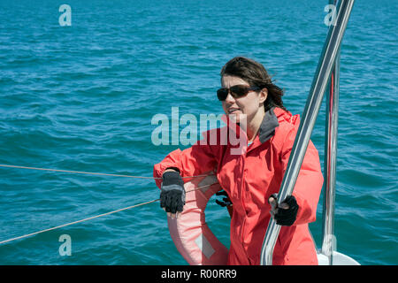 Smiling, young,  happy lady wearing sun glasses and dressed in red yachting jacker looking forward leaning against guard-rail wires on stern of yacht