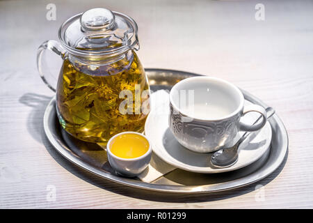 Hot linden-flower tea in transparent glass teapot, small cup of honey, and empty white porcelain cup and silver spoon on saucer arranged on silver tra