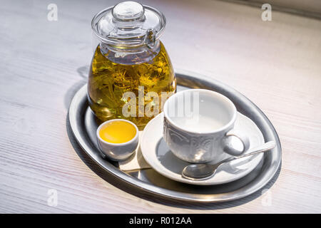 Hot linden-flower tea in transparent glass teapot, small cup of honey, and empty white porcelain cup and silver spoon on saucer arranged on silver tra