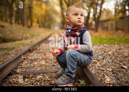 Little boy with biscuit in his hand sits on rails at railroad in autumn forest among yellow fallen leaves. Stock Photo