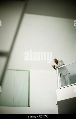 Mid-adult businessman leaning on a banister whilst texting on his cellphone in an office building. Stock Photo