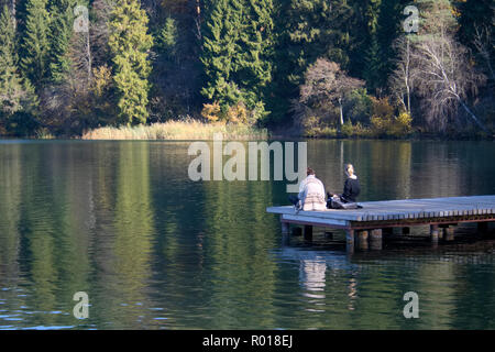 Two friends sitting on a wooden pier on a lake enjoying and relaxing the pure air of the forest Stock Photo
