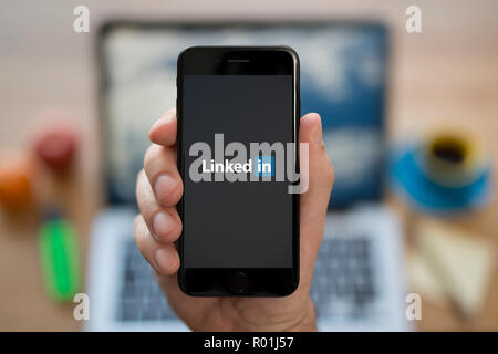 A man looks at his iPhone which displays the LinkedIn logo, while sat at his computer desk (Editorial use only). Stock Photo