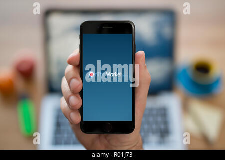 A man looks at his iPhone which displays the On The Market logo, while sat at his computer desk (Editorial use only). Stock Photo