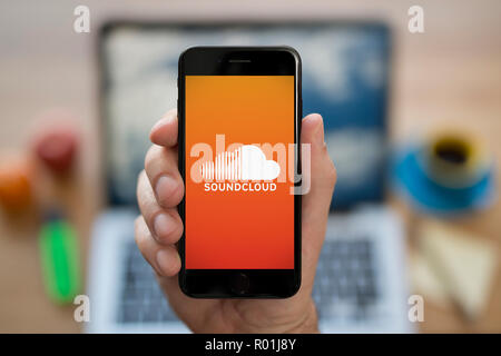 A man looks at his iPhone which displays the Soundcloud logo, while sat at his computer desk (Editorial use only). Stock Photo