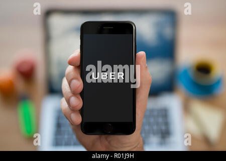A man looks at his iPhone which displays the Uber logo, while sat at his computer desk (Editorial use only). Stock Photo