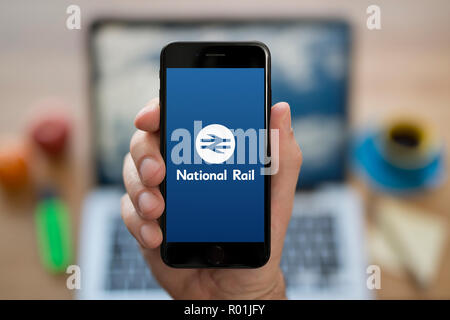 A man looks at his iPhone which displays the National Rail enquiries logo, while sat at his computer desk (Editorial use only). Stock Photo