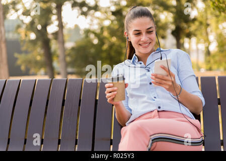 Young woman rests in autumn park on bench after a walk and listens to music on the phone at sunny day Stock Photo