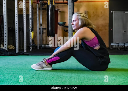 Young fit woman at the gym sitting on the ground and taking rest. Content female athlete at a fitness room taking a rest after workout