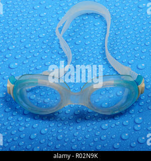 One single pair of white, blue and transparent swimming goggles lying on paper surface with water drops.  Water drops paper background. Stock Photo