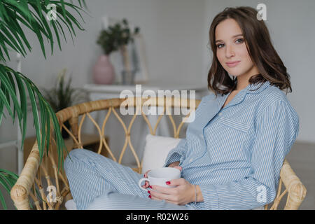 Sideways shot of pretty woman sits crossed legs in wicker chair, dressed in pyjamas, drinks hot coffee and latte, poses against cozy interior indoor,  Stock Photo