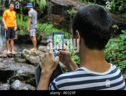 Selective focus view through the screen of mobile phone of teenagers in front of a scenic waterfall taking a group photo Stock Photo