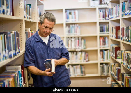 Mature man wearing reading glasses while reading the spine of a book inside a library. Stock Photo