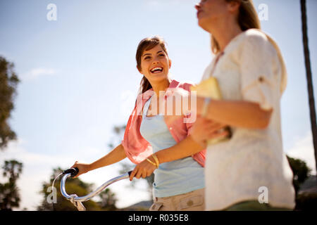 Two female friends walking through a park,  walking a bicycle. Stock Photo