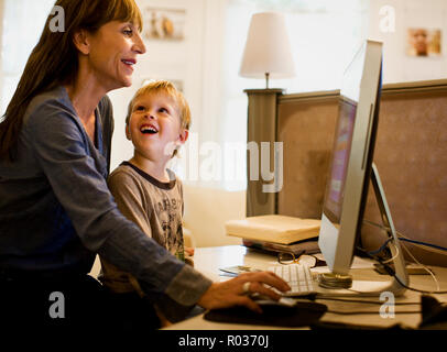 Young boy smiles up at his mother as they sit together at a home study in front of a large desktop computer screen. Stock Photo