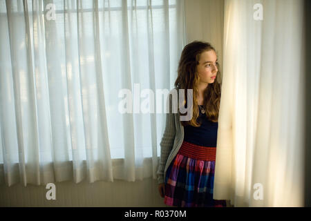 Girl looks pensivley out of a window from behind the edge of a curtain. Stock Photo