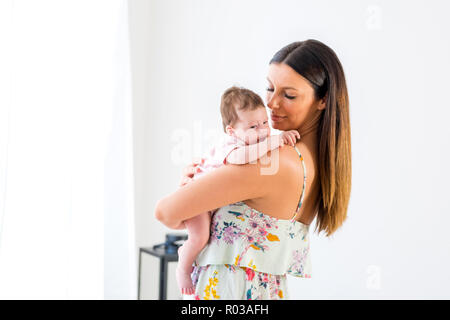 A beautiful young mother standing and holding her baby girl in her arms in front of a white wall. Stock Photo
