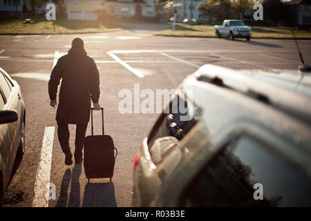 Mid adult man pulling a suitcase on wheels through a parking lot in the sunshine. Stock Photo