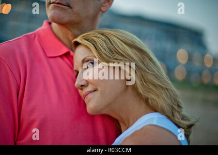 Woman lovingly resting her head on her husband's chest as they watch the sunset at the beach. Stock Photo