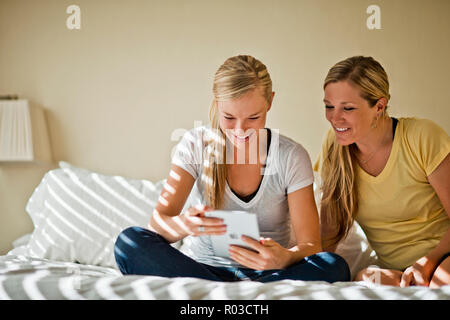 Two friends smile as they sit in a sunlit bedroom and watch something together on an electronic tablet. Stock Photo