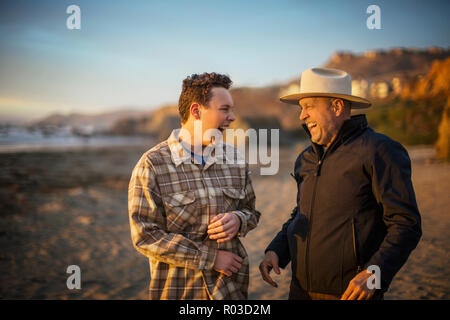 Teenage son and his middle-aged father laugh as they share a joke together as they walk on the beach at sunset. Stock Photo