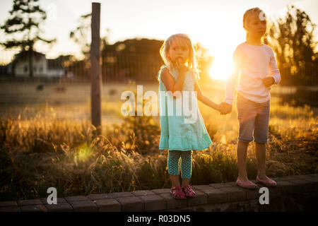 Two young blond sisters hold hands at the side of a country road at sunset. Stock Photo