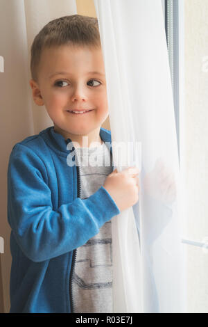 Portrait of a cute little Caucasian boy hiding behind a curtain next to the window Stock Photo